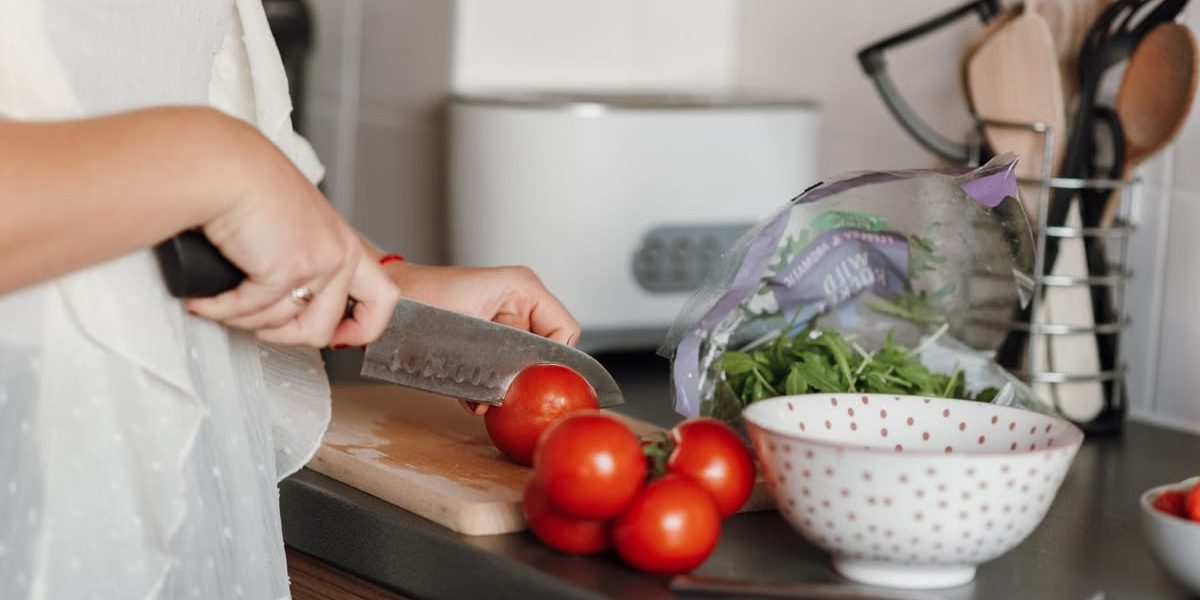 Crop anonymous housewife in casual clothes cutting fresh ripe tomatoes with sharp knife on wooden cutting board while cooking in kitchen