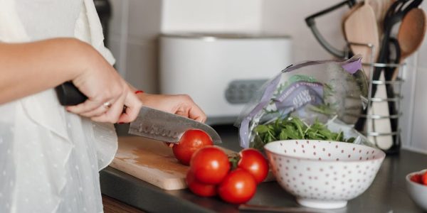 Crop anonymous housewife in casual clothes cutting fresh ripe tomatoes with sharp knife on wooden cutting board while cooking in kitchen